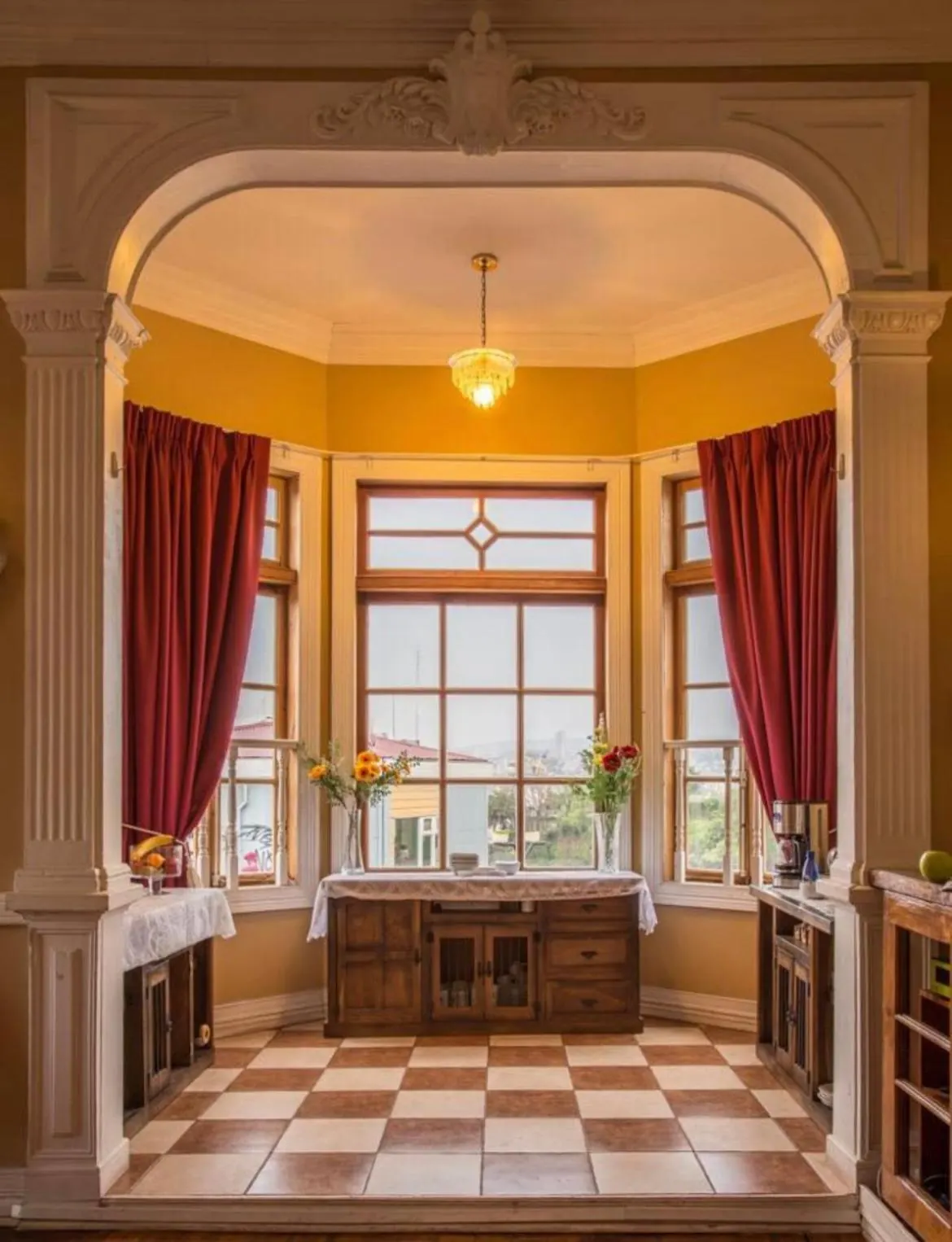 Un arco de molduras abre a un bow window con cortinas rojas, una cómoda con flores y piso de baldosas en damero.
