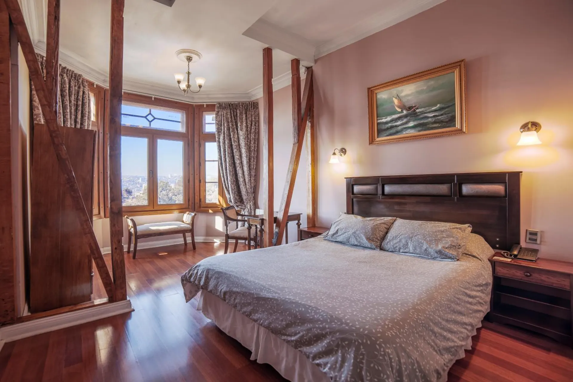 Habitación con vigas de madera a la vista, cama doble y al fondo un bow window con una banca bajo las ventanas.