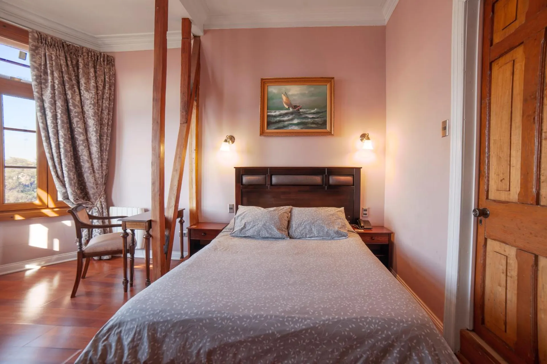 Cama doble bajo el cuadro de un velero, con vigas de madera, un escritorio con silla y la puerta de madera.