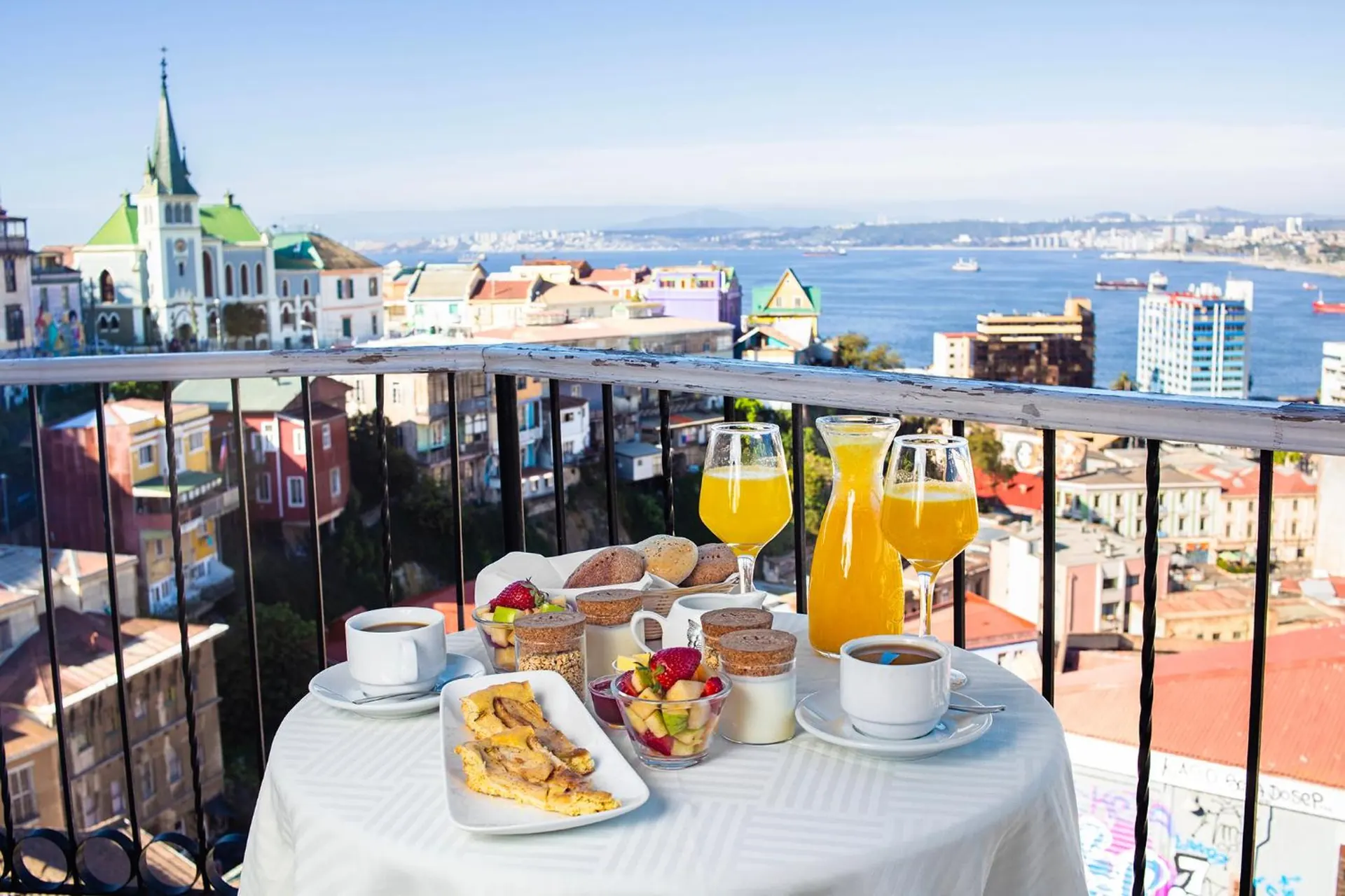 Un desayuno servido en un balcón: jugo de naranja, café, frutas y huevos, con la iglesia y la bahía de fondo.