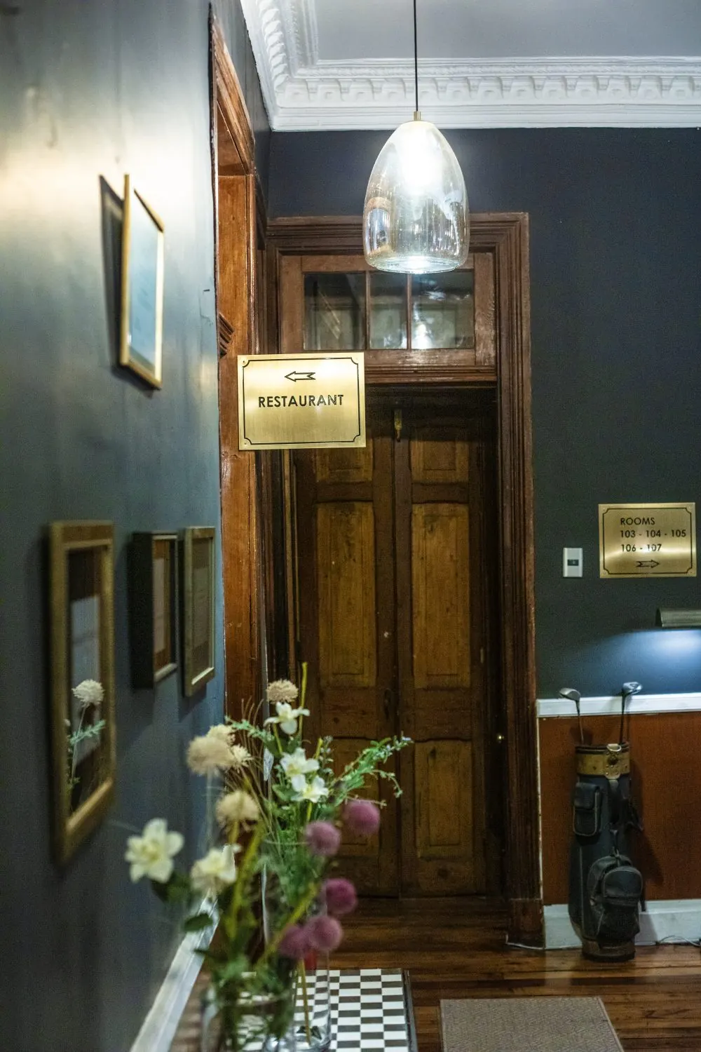 Un pasillo de muros oscuros con un letrero de bronce que dice Restaurant, una puerta de madera y un florero.