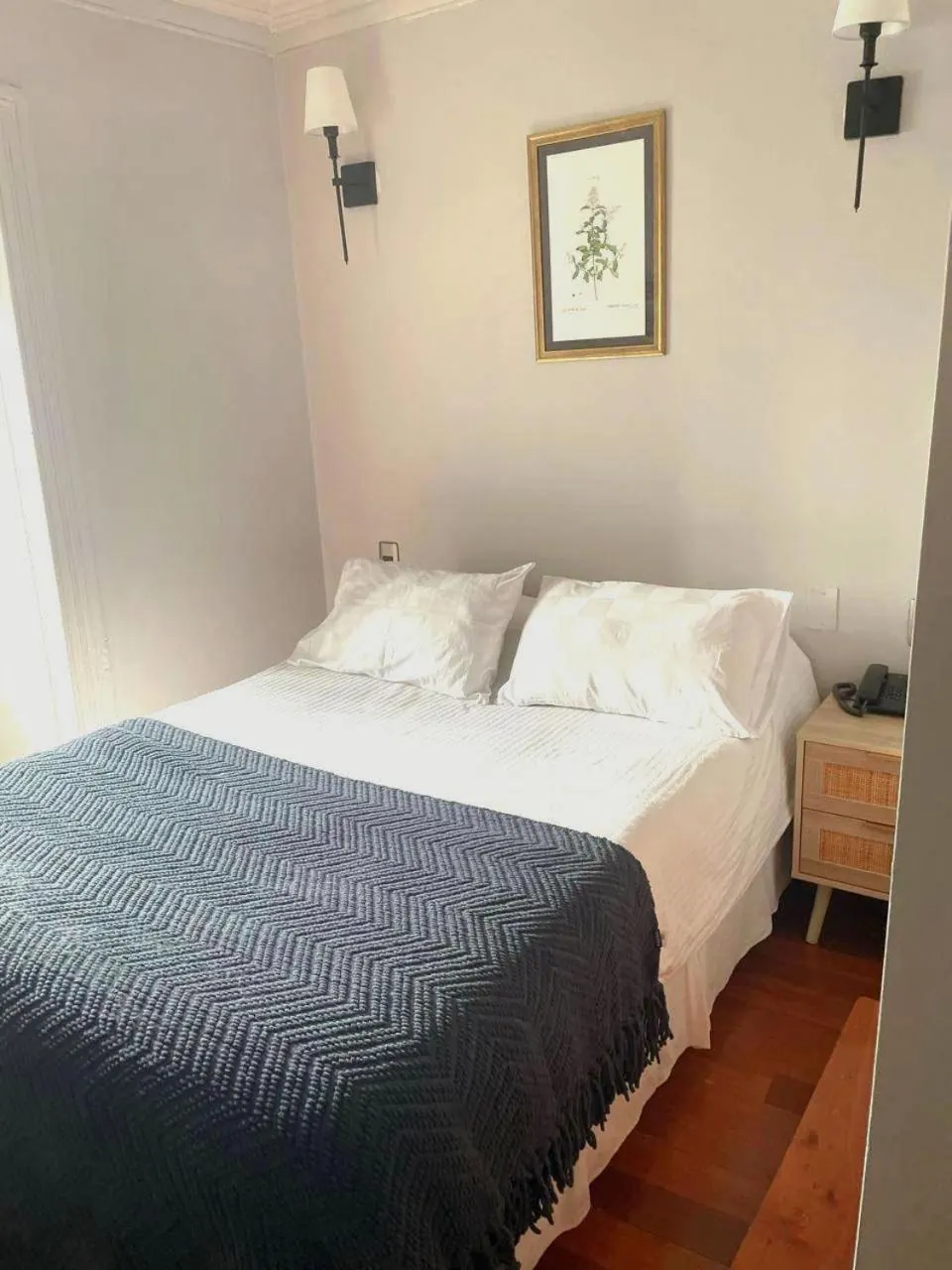 La Single: cama con cubrecama azul tejido, muros claros, un cuadro botánico y la ventana a la derecha.