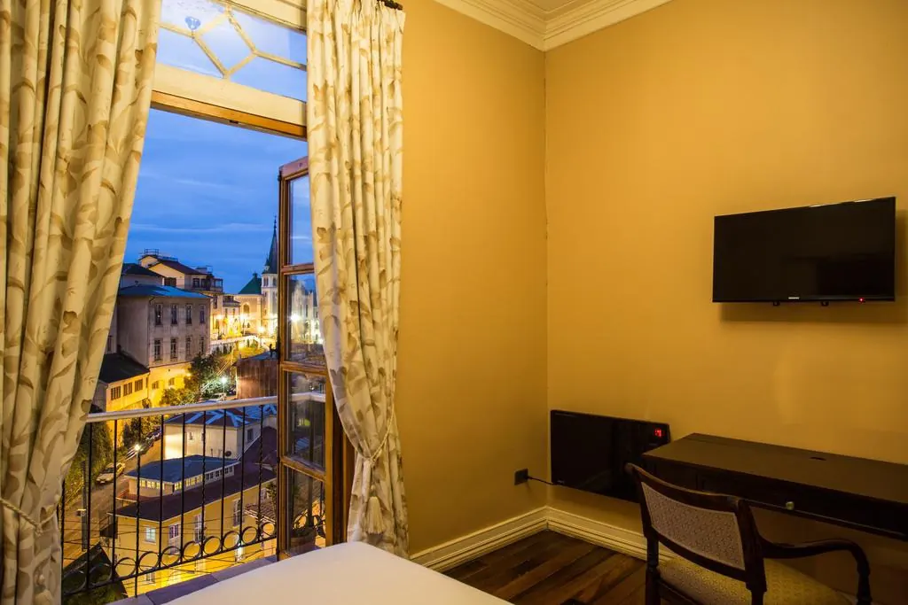 Habitación de muros amarillos con la ventana abierta al balcón y al cerro iluminado de noche.