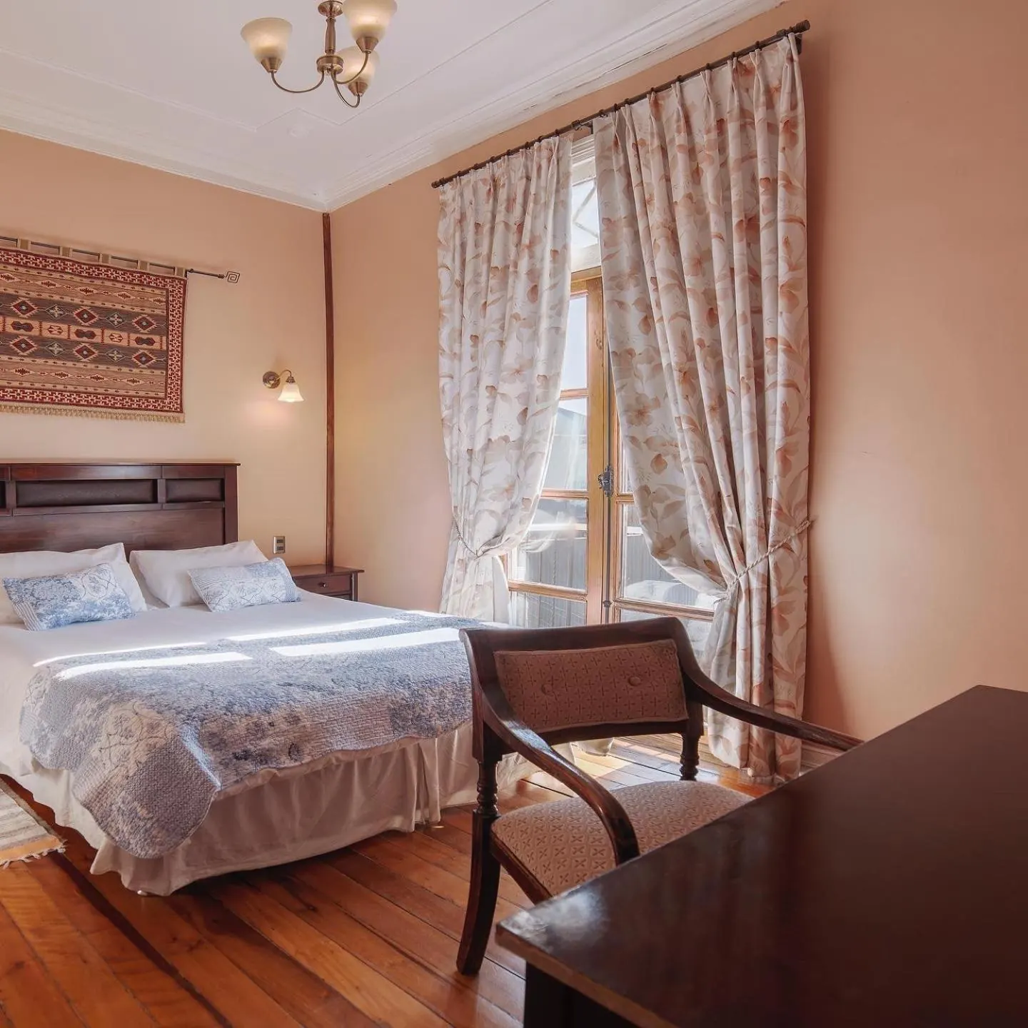 Habitación rosada con un tapiz sobre la cama, un escritorio con silla de madera y cortinas de flores.