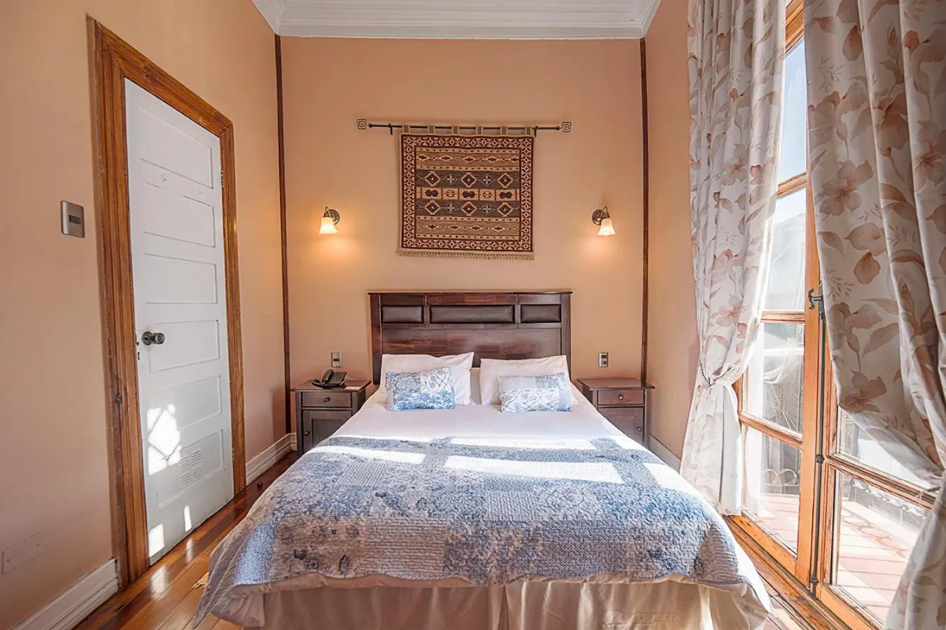 Cama doble con cubrecama azul y blanco bajo un tapiz, con la ventana del balcón abierta.
