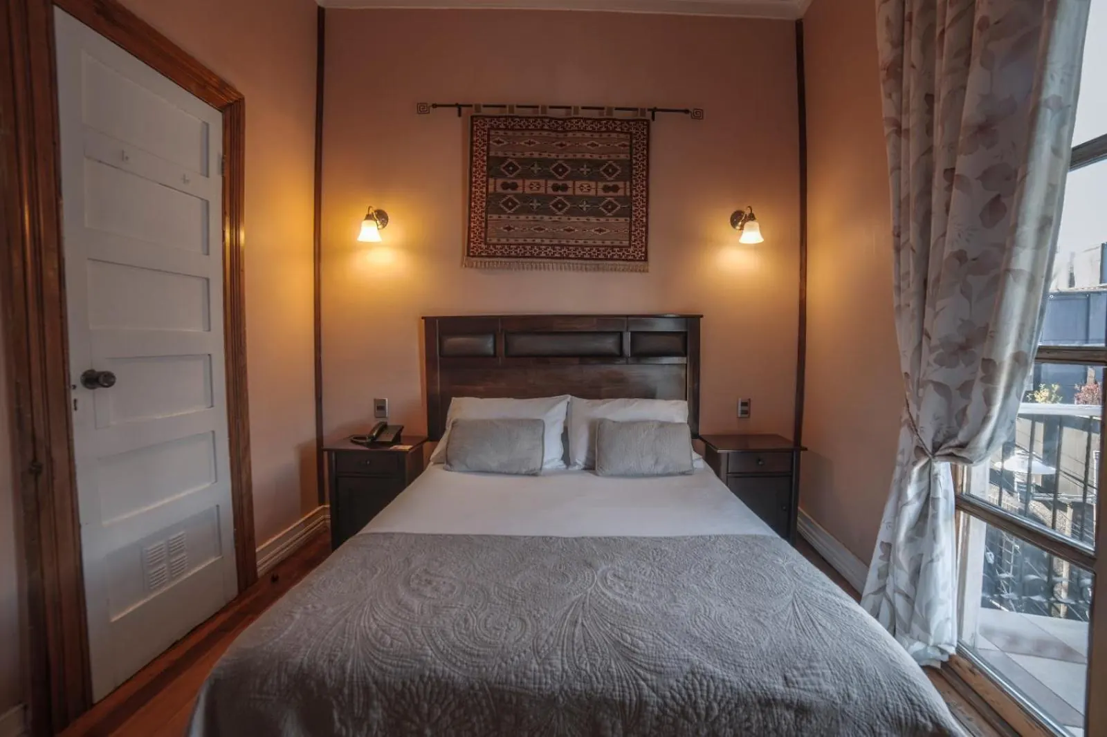 Cama doble bajo un tapiz tejido, con lámparas de pared, una puerta blanca y la ventana con cortinas.