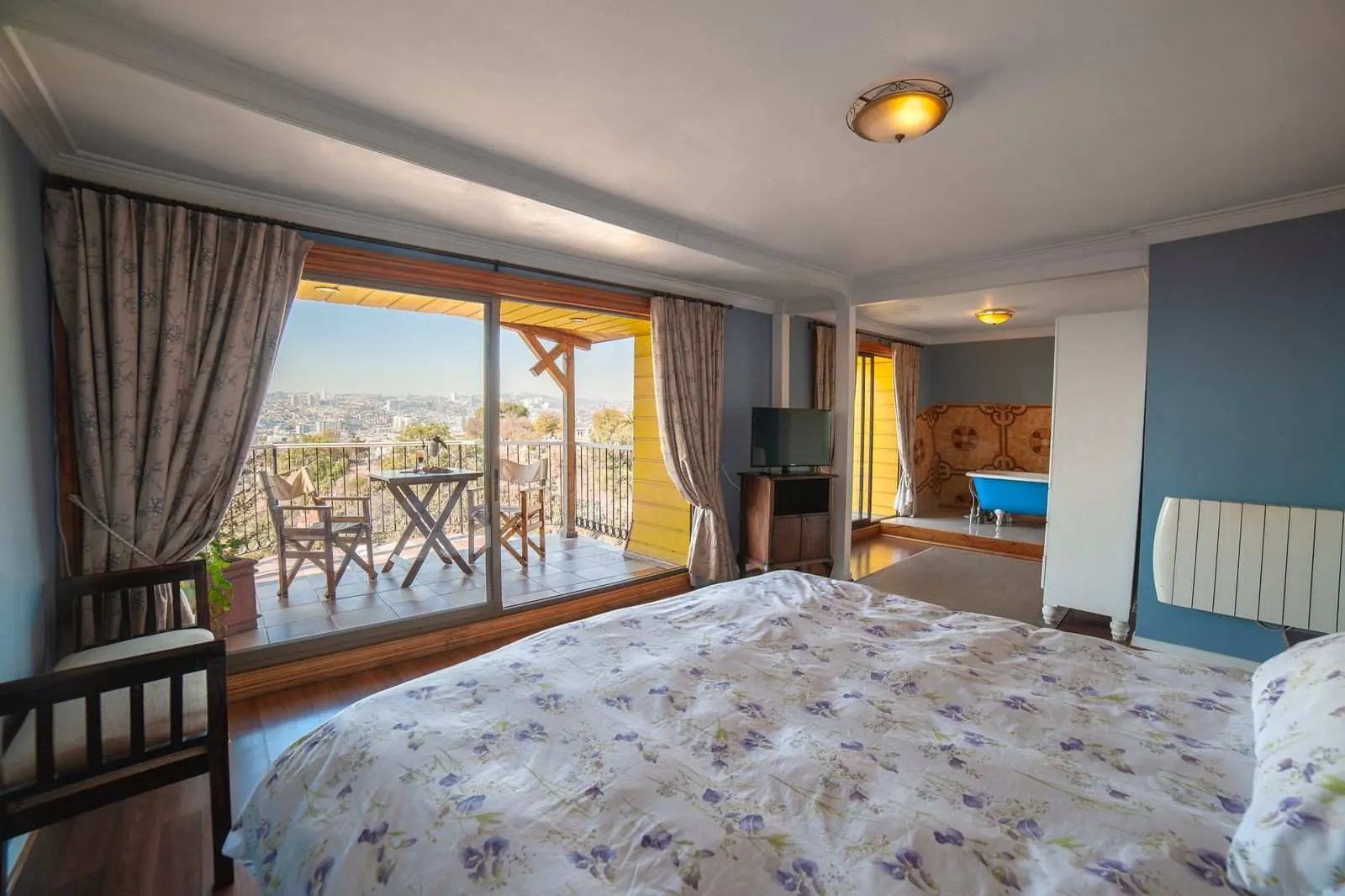La Suite: cama con cubrecama de flores lilas, puertas de vidrio abiertas a la terraza con mesa y sillas, y al fondo la tina azul.