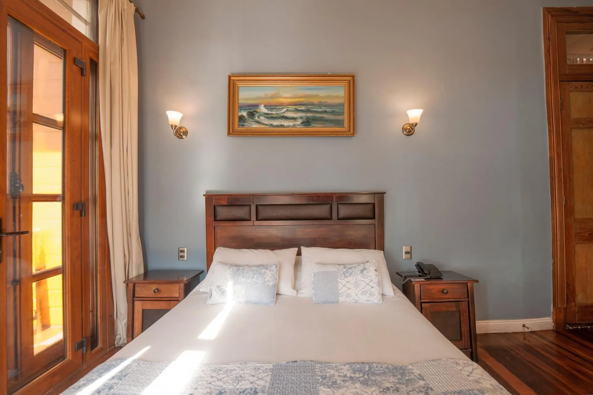 Cama de respaldo de madera bajo un cuadro de un velero, muros celestes y una puerta de madera abierta.