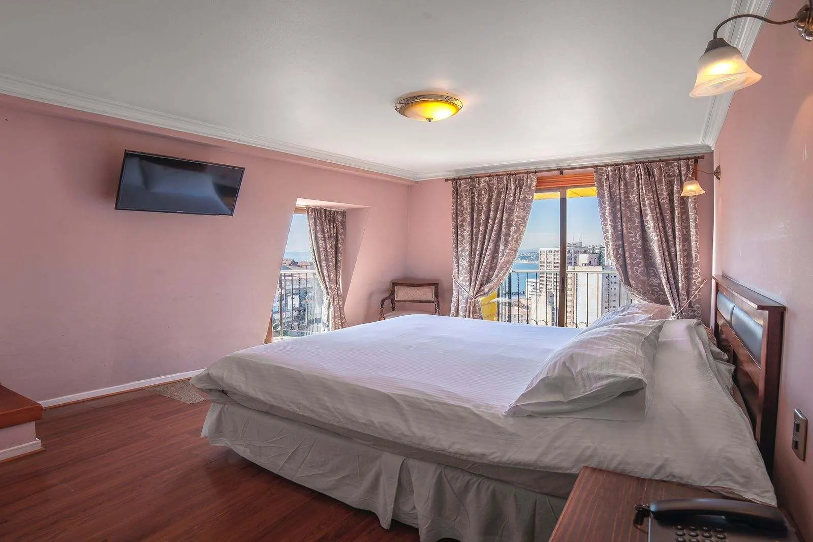 Habitación de muros rosados con cama doble, televisor en la pared y ventanales con cortinas que miran a la ciudad.