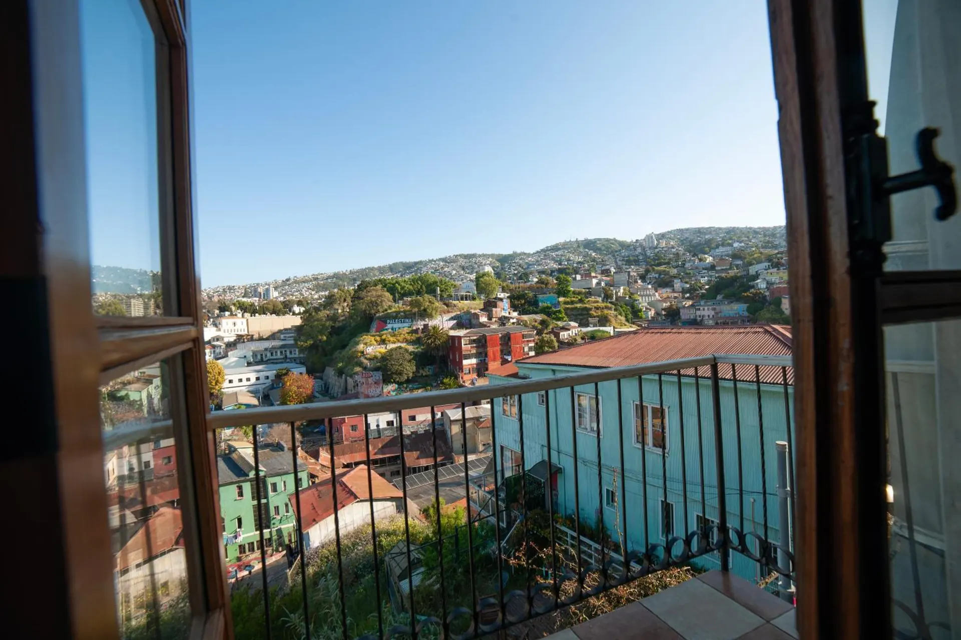 Una ventana abierta sobre un balcón de fierro, con las casas de colores de los cerros de Valparaíso enfrente.